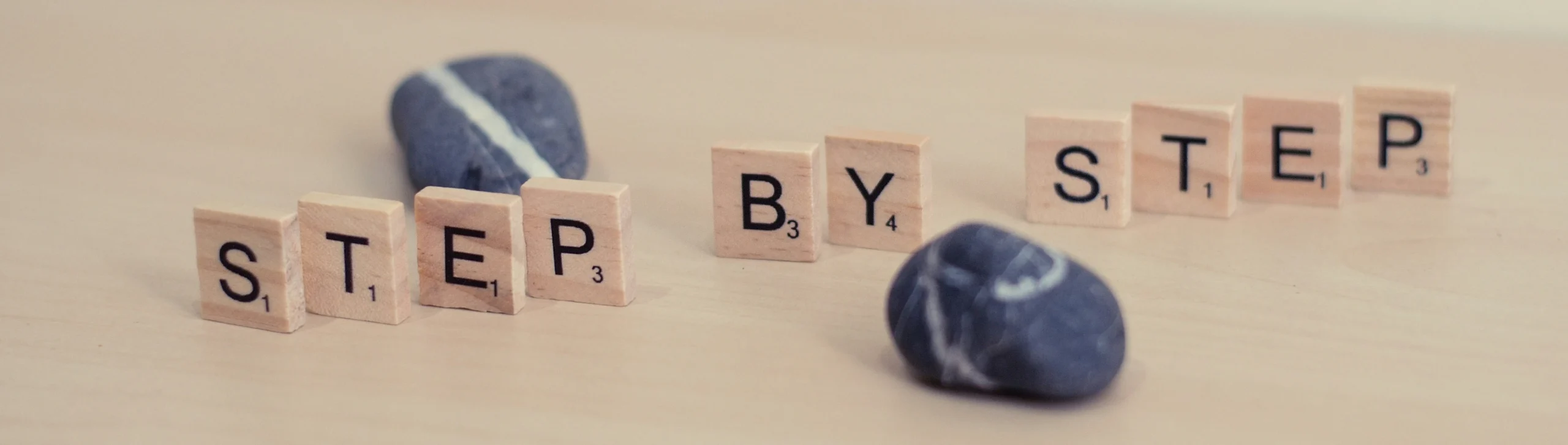Wooden letter tiles spell 'STEP BY STEP' on a light wood surface with two dark stones nearby.
